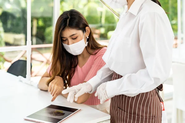 Asian waitress wear face mask and face shield using tablet to show restaurant electronic menu and recommend to customer. New normal technology and lifestyle food and dining concept.