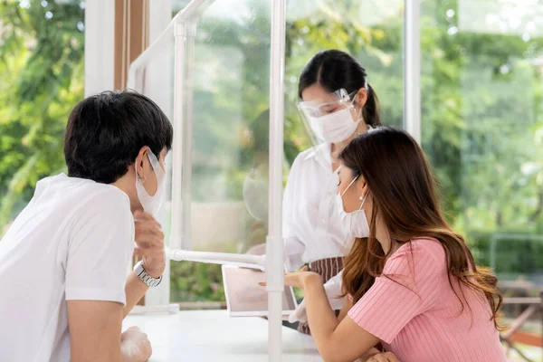 Asian waitress wear face mask and face shield using tablet to show restaurant electronic menu and recommend to couple customers. New normal technology and lifestyle food and dining concept.