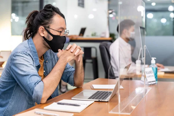 Asian office employee businessman wear protective face mask work in new normal office with interracial colleague in background as social distance practice prevent coronavirus COVID-19.