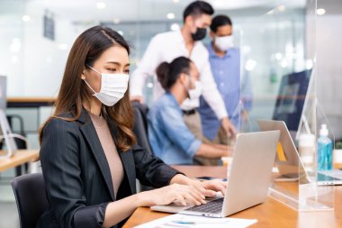 Asyalı ofis çalışanı iş kadını portresi yeni normal ofiste koruyucu yüz maskesi takıyor ve sosyal uzaklık uygulaması Coronavirus COVID-19 'u engellerken ırklar arası takım arka planda yer alıyor..