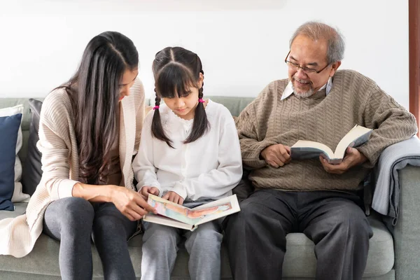 Asian cute girl reading story cartoon book with her mom in living room with grandfather reading fiction book in background. Happiness multigenerational family and domestic lifestyle concept.