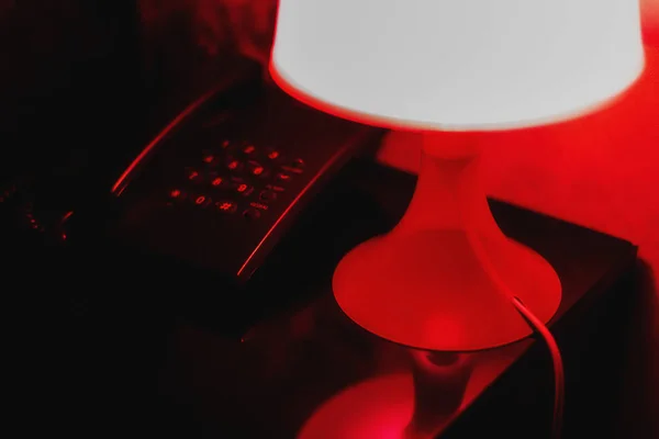 De nacht lamp staat op het nachtkastje naast de telefoon en de kamer verlicht met rood licht het creëren van een intieme sfeer in het Motel kamer — Stockfoto