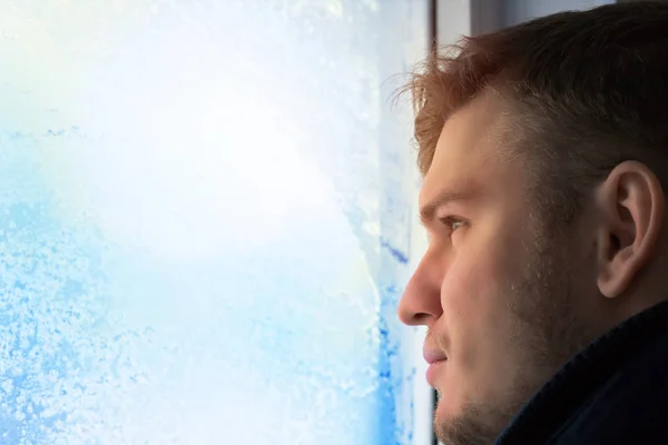 Joven hombre atractivo en el día de invierno mira por la ventana. Vidrio en hoarfrost y copos de nieve. Retrato de primer plano en luz natural . —  Fotos de Stock