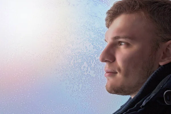 White attractive man in profile close - up against the window in the frost. Soft sunlight through the glass illuminates the persons face Winter theme.