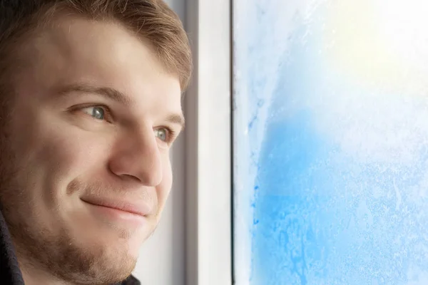 Un joven guapo mira la ventana congelada en la helada y sonríe. Buen humor en previsión de las vacaciones y el fin de semana. Retrato de un tipo con luz sin afeitar primer plano — Foto de Stock