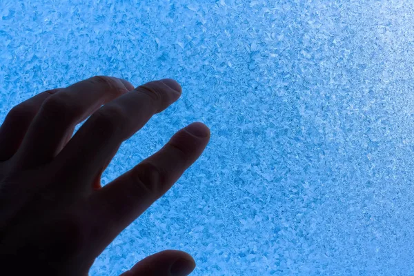Silhouette einer Menschenhand auf dem Hintergrund eines eisblauen Fensters. Frost und schwacher Schein der Sonne durch das Glas. Kopierraum rechts. — Stockfoto
