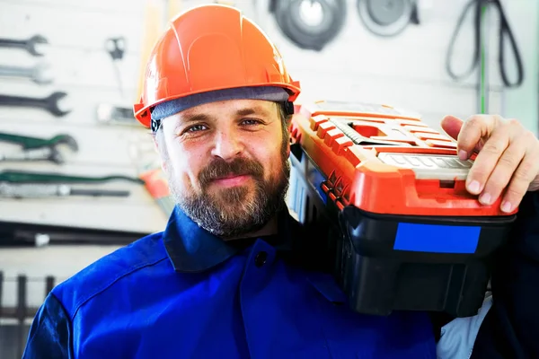 Bir işçi bir kask omzunun üstünde alet kutusu içinde doğrudan içine belgili tanımlık fotoğraf makinesi ve gülümsüyor görünüyor. Sakallı adam iş elbiseleri araçları ile duvar arka plan üzerinde beyaz görünüm.