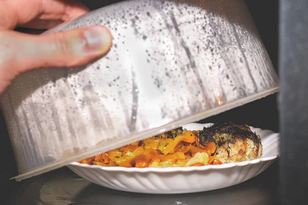 Tányér étellel melegíteni a mikrohullámú sütőben. Előnyei és veszélyei sugárzás mikrohullámok az élelmiszerek kérdését. Szakács megnyitja a műanyag fedelet, és láthatjuk az ebéd a tányéron. — Stock Fotó
