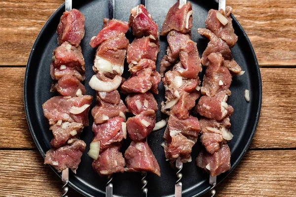 A number of skewers with meat laid out in a black plate. Close-up of mouth-watering pieces of beef ready to fry. Meat dish. Top view on wooden background — Stock Photo, Image
