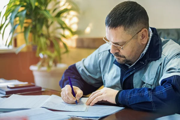En manlig ingenjör i glas på kontoret vid bordet undertecknar dokumenten. Inspektion, registrering av recept och kränkningar i loggen. — Stockfoto