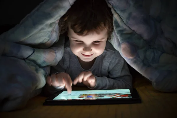 小女孩在床上看她的平板电脑 从设备屏幕上照亮微笑的孩子脸 孩子们穿着睡衣在被窝里拿着一块平板电脑 夜间时间 — 图库照片