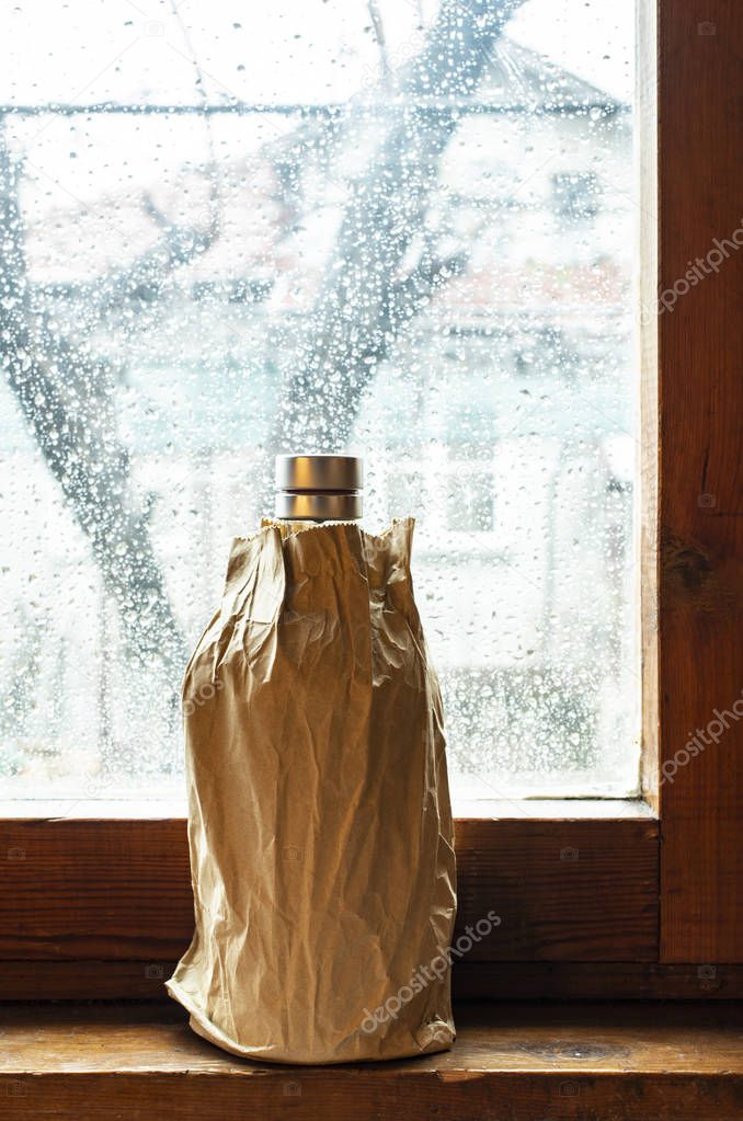 Bottle with alcohol in a brown pack paper placed to the window at rainy day. Wrinkled paper package. Hidden bottle alcohol in paper. Concept for stealthily drinking and alcoholism.