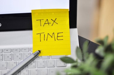 yellow paper sticker with text: tax time, workplace table with laptop and pen 