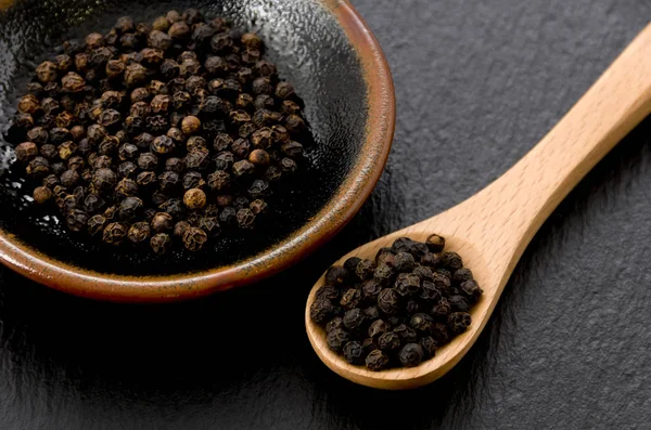 Black pepper in black dish with wooden spoon on black stone plate