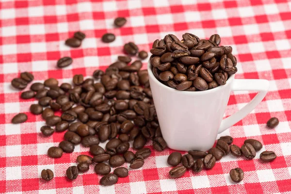 A white coffee mug is  filled with brown coffee beans. The coffee beans are also spread out on the kitchen table with a red checkered tablecloth as a background.