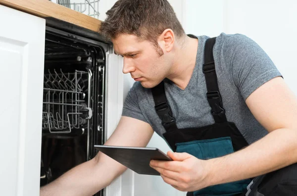 Handyman with tablet pc repairing domestic dishwasher in the kitchen.