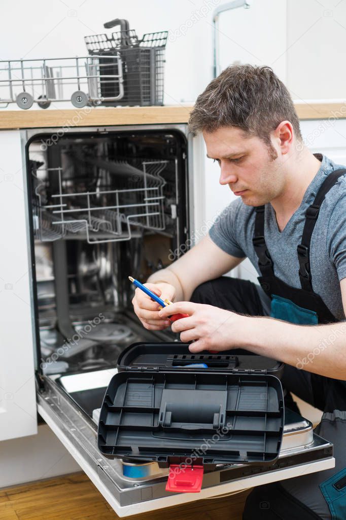 Professional handyman in overalls repairing domestic dishwasher in the kitchen.