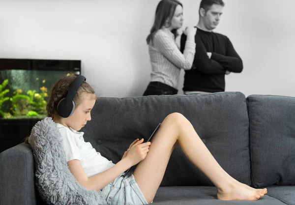 Liten flicka i hörlurarna med Tablet PC medan hennes föräldrar i gräl i bakgrunden. — Stockfoto