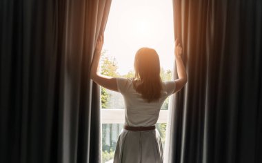 Güneşli sabah otel odasında perdeleri açan kadın.