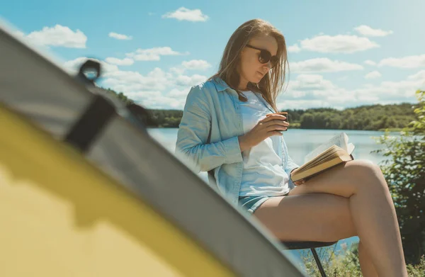 Woman reading a book near camping tent by the lake.