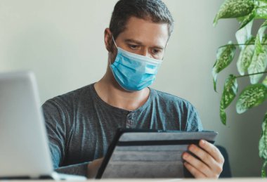 Tıp maskeli bir adam ofiste tablet bilgisayar kullanıyor..