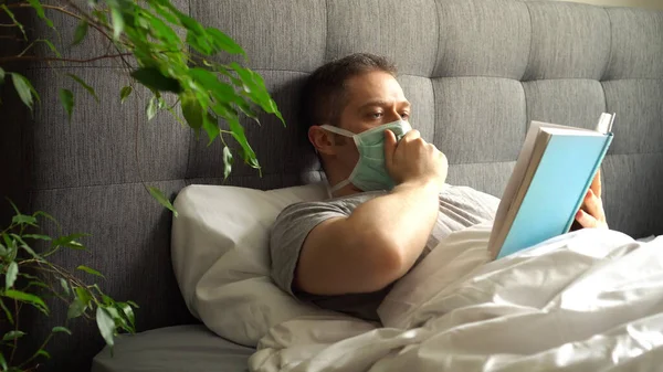 ベッドで医学マスク読書本を持つ病気の男 ホーム隔離の概念 — ストック写真