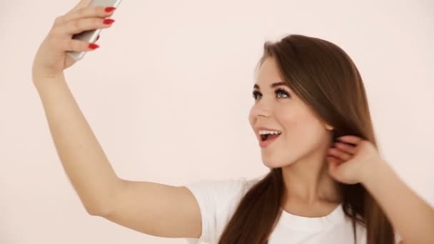 Juventud moderna. hermosa chica de pelo largo de aspecto europeo con el pelo castaño hace selfie en un teléfono inteligente cerca de una pared blanca — Vídeos de Stock