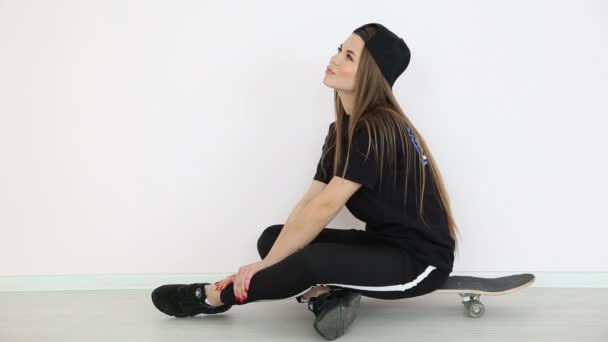 Девушка-подросток в модной хип-хоп одежде и кепке позирует на белой стене со скейтбордом — стоковое видео