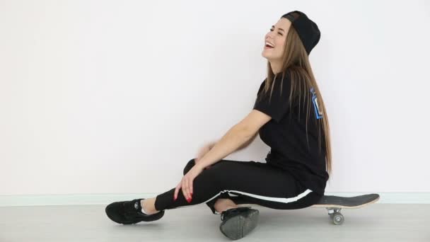 Девушка-подросток в модной хип-хоп одежде и кепке позирует на белой стене со скейтбордом — стоковое видео