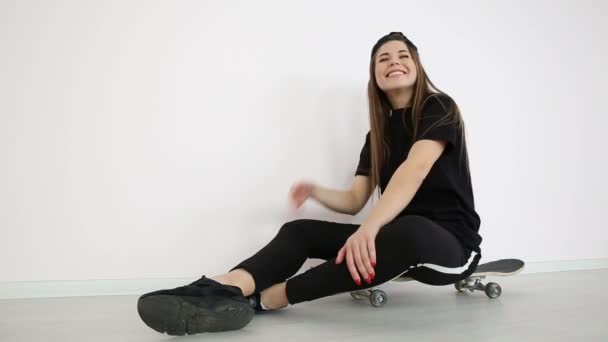 Teenage girl in trendy hip hop clothes and cap posing against white wall with skateboard — Stock Video