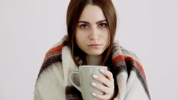 A beautiful cold girl of Eurarean appearance in a woolen blanket is drinking hot tea by the white wall. — Stock Video