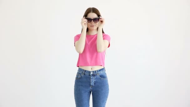 Belle fille gaie dans les lunettes de soleil, haut rose et jeans posant contre le mur blanc — Video