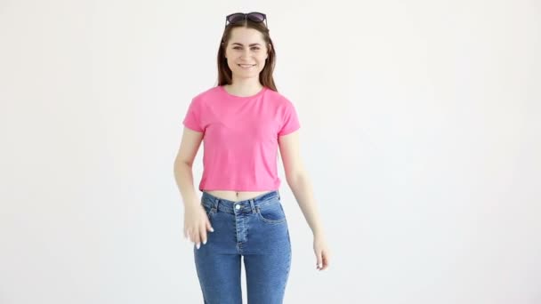 Belle fille gaie dans les lunettes de soleil, haut rose et jeans posant contre le mur blanc — Video