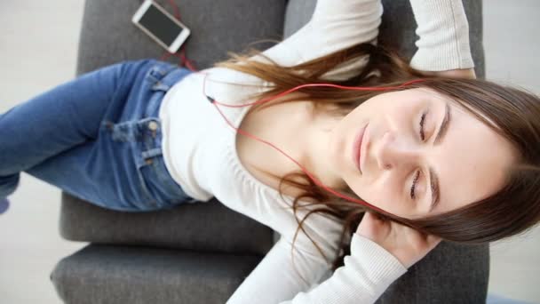 Vacker flicka lyssnar på musik med mobiltelefon i hörlurar på soffan hemma — Stockvideo