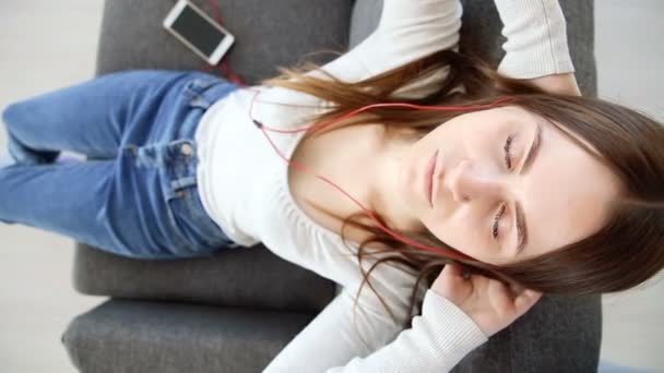 Красивая девушка слушает музыку с мобильного телефона в наушниках на диване дома — стоковое видео