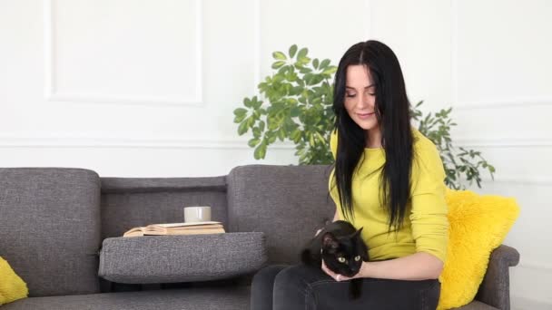 Η αγάπη για τα κατοικίδια ζώα. Όμορφη σκοτάδι μαλλιά κορίτσι με μια μαύρη γάτα είναι να χαλαρώσετε στο σπίτι στο σαλόνι στον καναπέ. — Αρχείο Βίντεο
