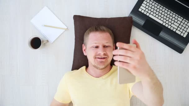 Hombre-freelancer hablando en el teléfono inteligente de comunicación de vídeo en casa en el suelo cerca de la computadora portátil — Vídeos de Stock