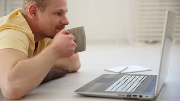 Une pause dans le travail d'un pigiste - un homme parlant à ses collègues dans un chat vidéo sur un ordinateur portable sur le sol et buvant du café — Video