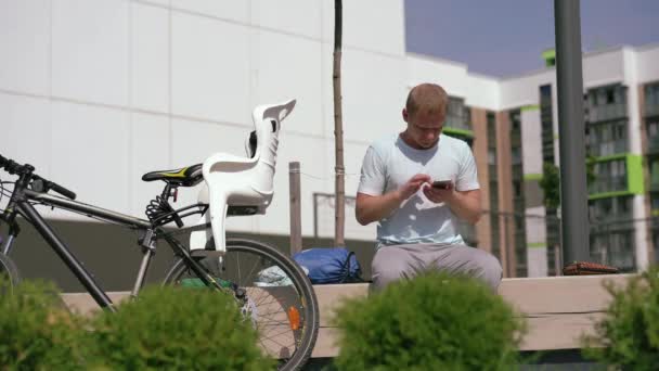 Современная жизнь в большом мире - молодой человек сидит в городе на скамейке и переписывает в смартфоне — стоковое видео