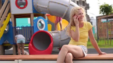 modern bir şehir - hayatta rahat kıyafetler kıvırcık saçlı kız bir smartphone oyun alanında yakınındaki bir bankta oturur