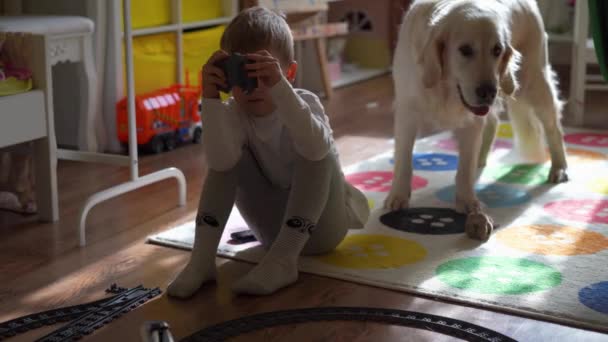 Style de vie maison. un petit garçon joue dans la chambre des enfants avec un train de jouets, un gros chien le regarde — Video