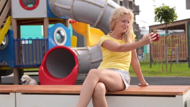 Vida em uma cidade moderna - menina com cabelo encaracolado em roupas casuais senta-se em um banco perto do parque infantil com um smartphone — Vídeo de Stock