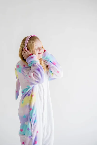 Modern fashion - beautiful blonde girl listens to music with headphones on a white background in kigurumi pajamas — Stock Photo, Image