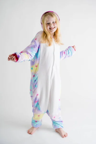 Moderní móda - krásná blond dívka poslouchá hudbu se sluchátky na bílém pozadí v kigurumi pyžama — Stock fotografie