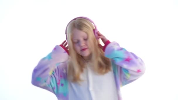 モダンなファッション - ヘッドフォンと着ぐるみパジャマ - 白で隔離の白い背景の上でダンス音楽を聴いて美しいブロンドの女の子 — ストック動画