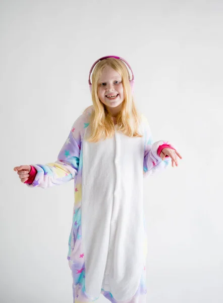 Moderní móda - krásná blond dívka poslouchá hudbu se sluchátky na bílém pozadí v kigurumi pyžama — Stock fotografie