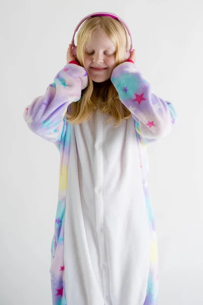 Moderní móda - krásná blond dívka poslouchá hudbu se sluchátky na bílém pozadí v kigurumi pyžama — Stock fotografie