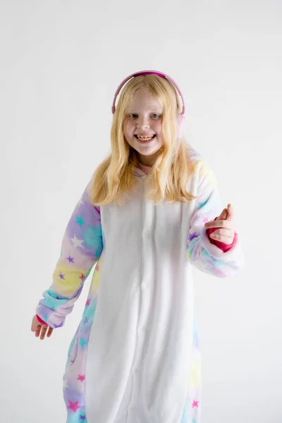 Moderní móda - krásná blond dívka poslouchá hudbu se sluchátky na bílém pozadí v kigurumi pyžama — Stock fotografie