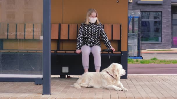 Pandemia koronawirusów w mieście. dziewczyna w masce ochronnej siedzi na przystanku komunikacji miejskiej z psem — Wideo stockowe