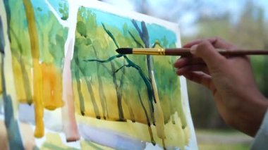 Ressamın eli açık havada manzarayı suluboya boyuyor.